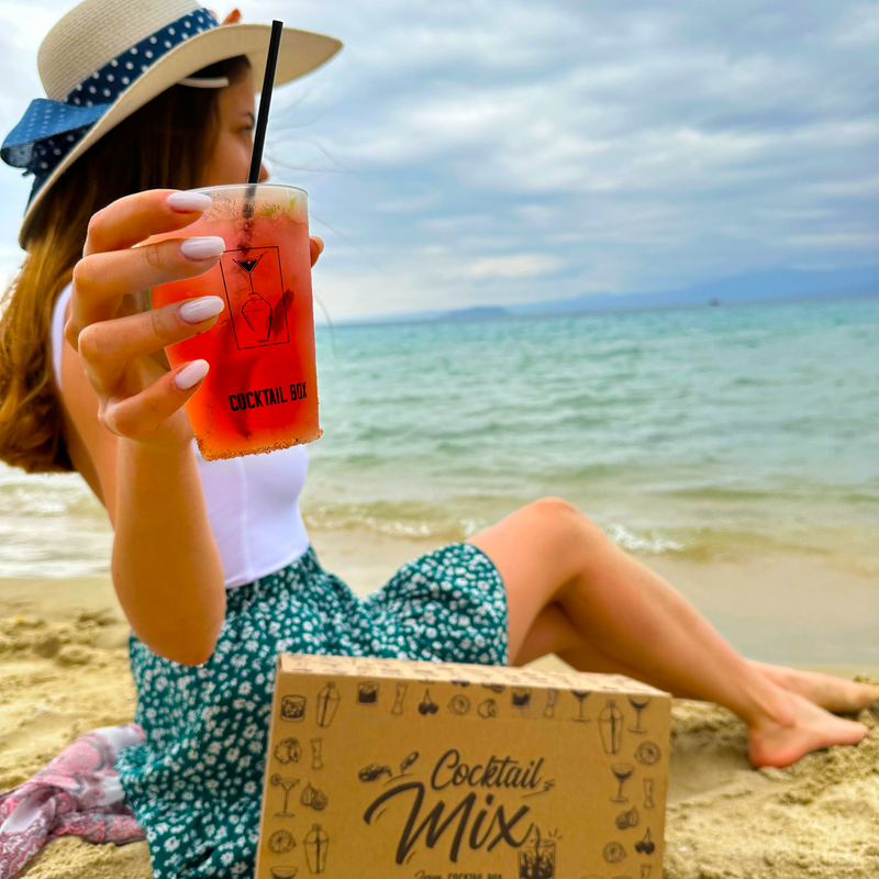 Жена на плажа с червен коктейл в брандирана чаша CocktailBox и кутия Cocktail Mix в пясъка