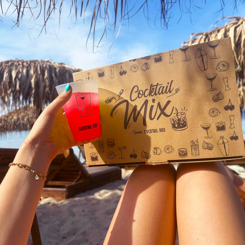 Кутия Cocktail Mix и червен коктейл в ръка на шезлонг на плажа