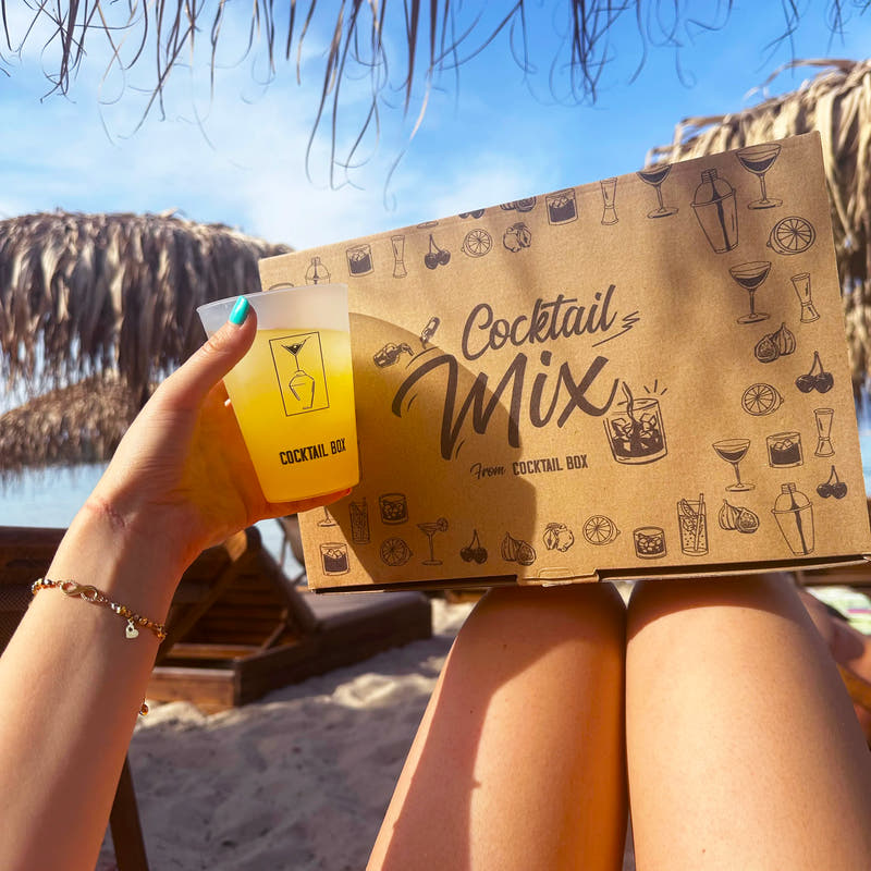 Кутия Cocktail Mix и жълт коктейл в ръка на шезлонг на плажа