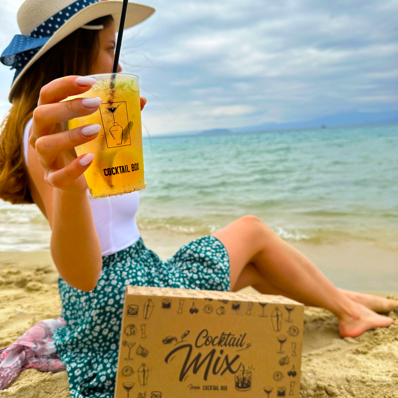 Жена на плажа с жълт коктейл в брандирана чаша CocktailBox и кутия Cocktail Mix в пясъка